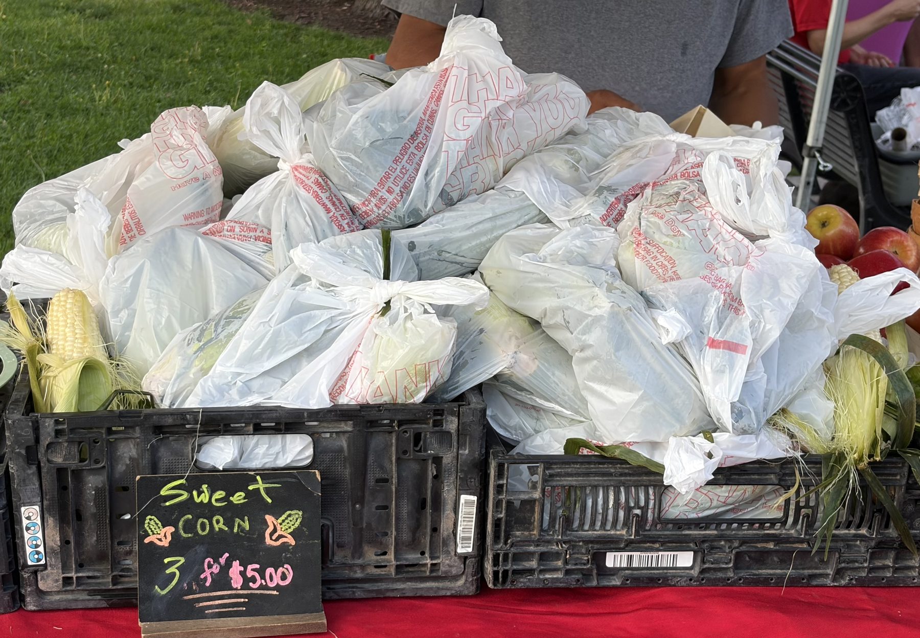 Bagged sweet corn heaped in black crates, with a chalkboard reading Sweet Corn 3 for $5.00