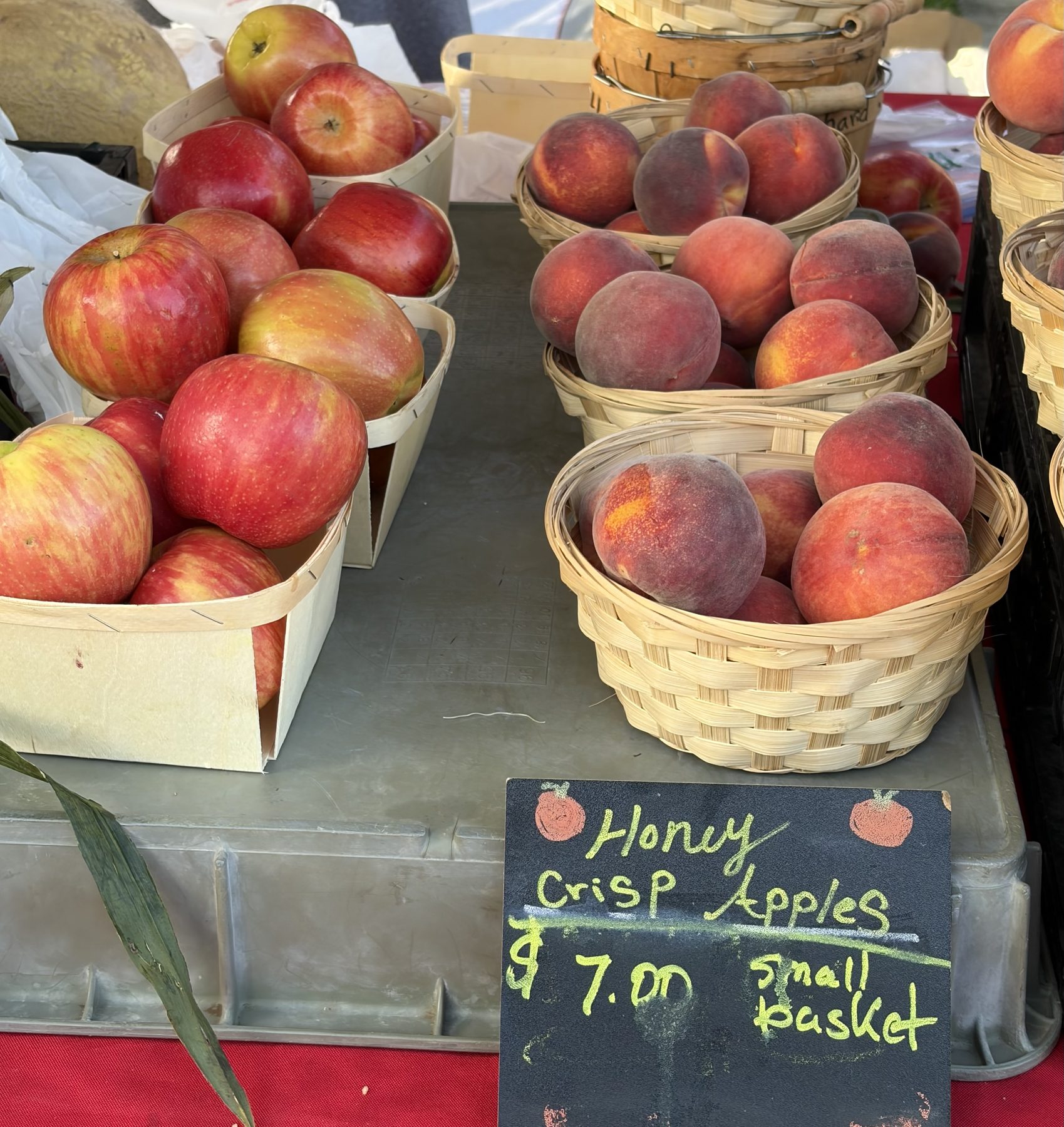 Wooden baskets of Honey Crisp apples beside baskets of peaches, with a chalkboard reading Honey Crisp Apples $7.00 small basket