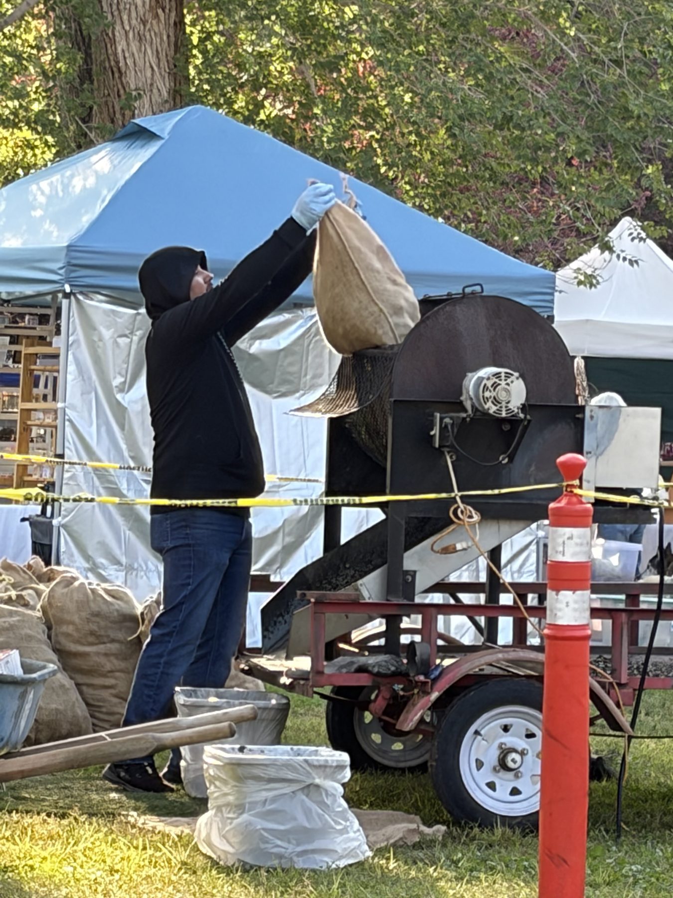 A worker in a wide-brimmed hat holding a burlap sack at the drum chile roaster, with the roaster trailer behind