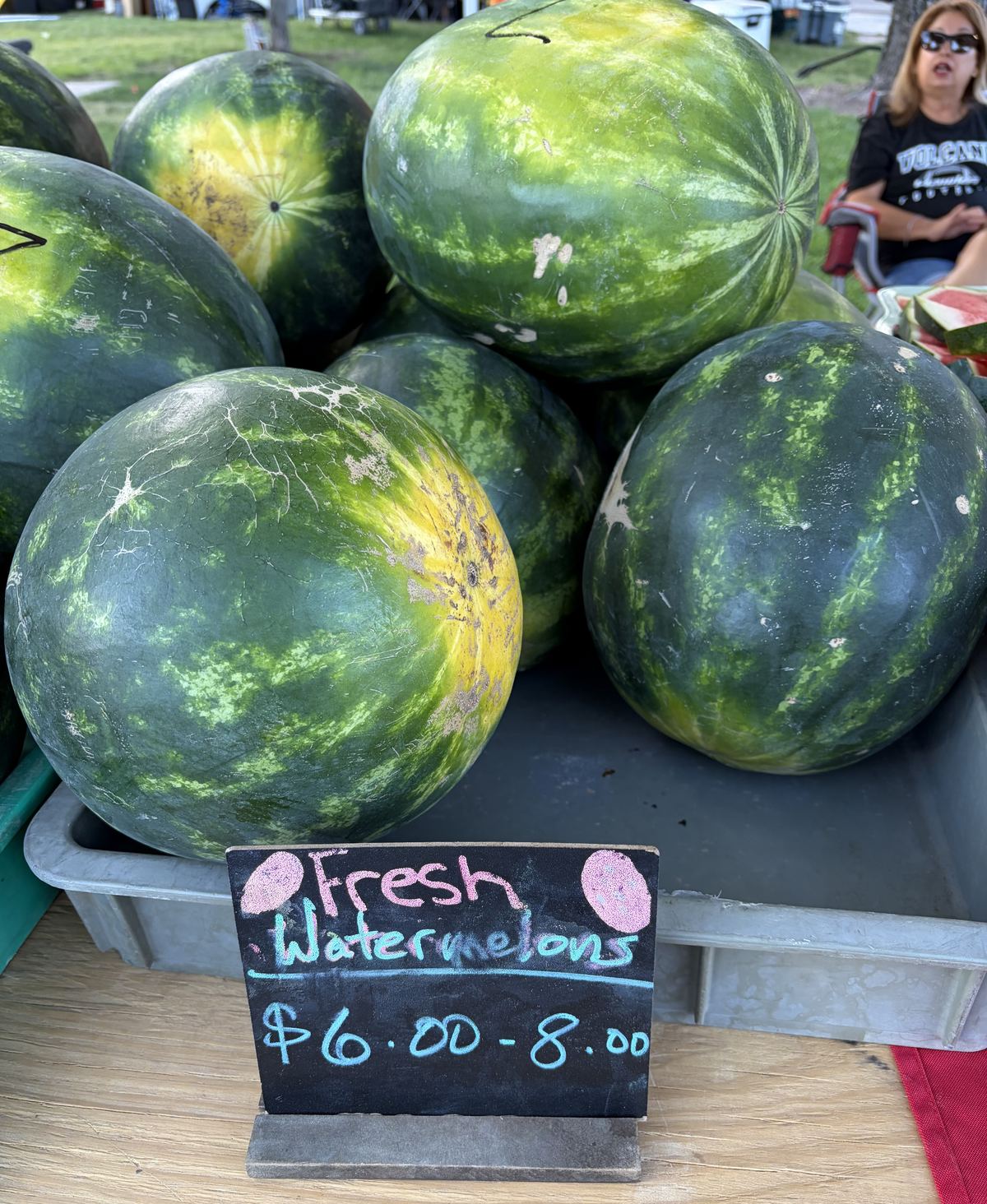 Stacked whole watermelons beside a chalkboard reading Fresh Watermelons $6.00-8.00